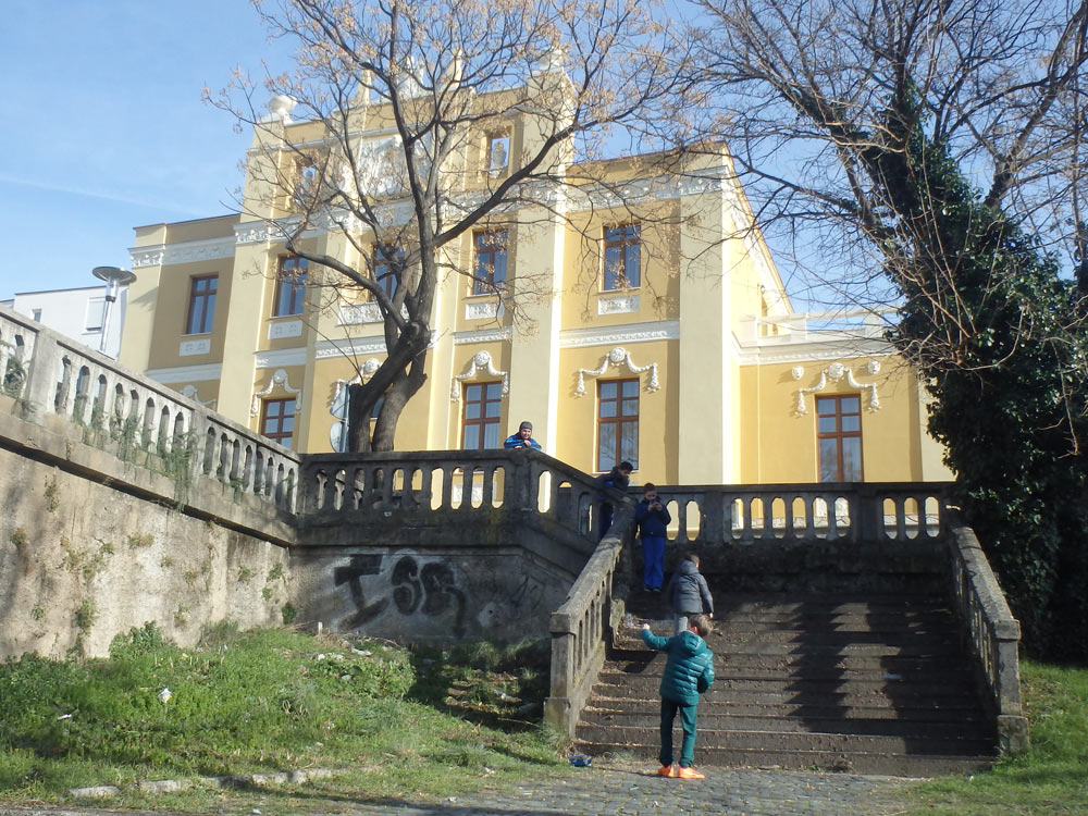 Kids setting off cherry bombs in front of a big yellow building in the hills overlooking the river in Mostar.